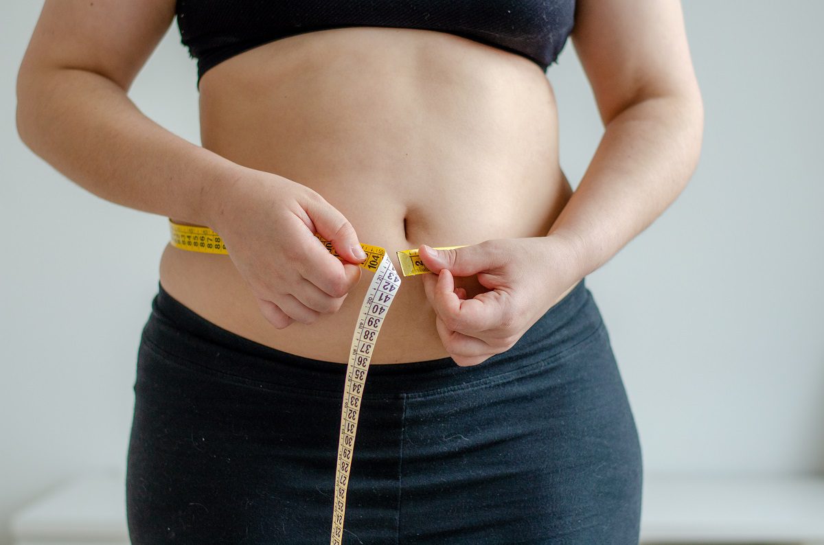 A woman measures her stomach with a roll of measuring tape.